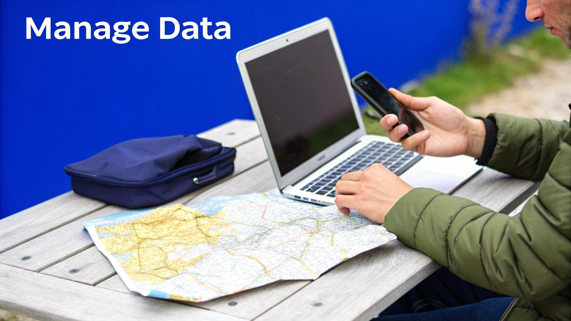 Person working outdoors on laptop and phone with a map, managing data on a wooden table.